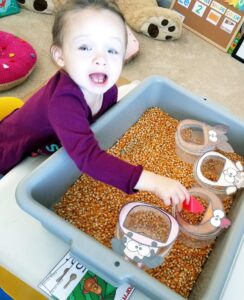 Toddler girl playing in spring theme sensory bin with corn and containers with farm animal faces to feed the corn.