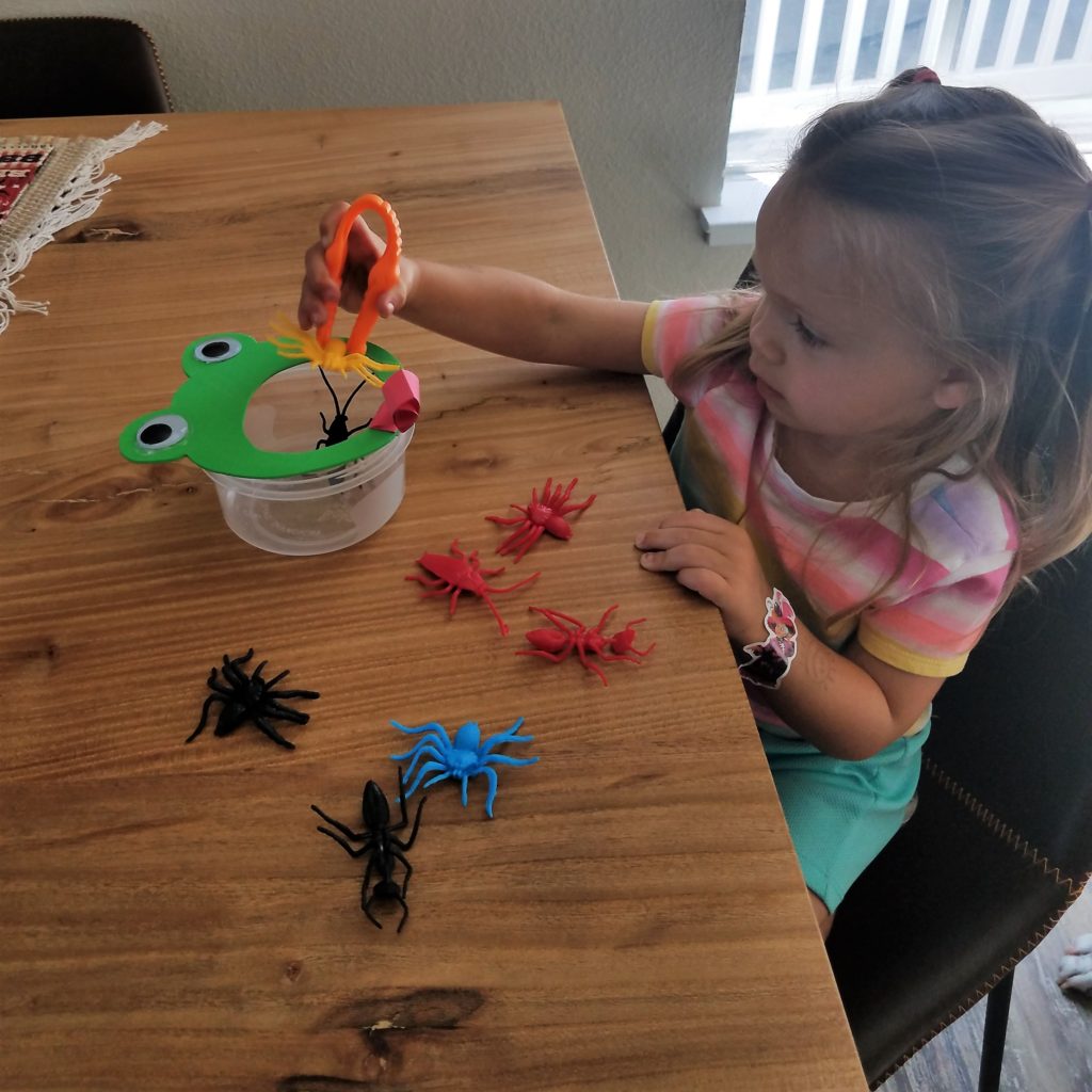 Spring theme Preschool activity with girl sitting at table with container with frog face and mouth cut out using tweezers to feed the frog fake bugs.