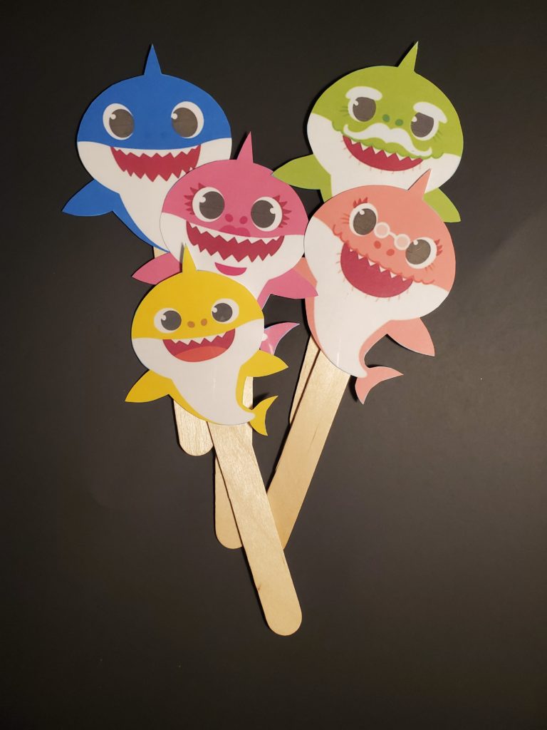 5 paper cut outs of colorful cartoon sharks in blue, green. pink, orange and yellow, glued to the top of popsicle sticks.