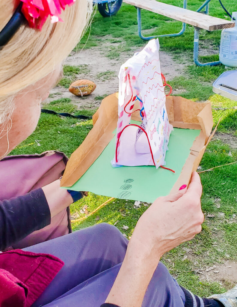 Dog house made with paper, pipe cleaners, tape, straws and string on a green piece of paper with a low brown paper bag fence