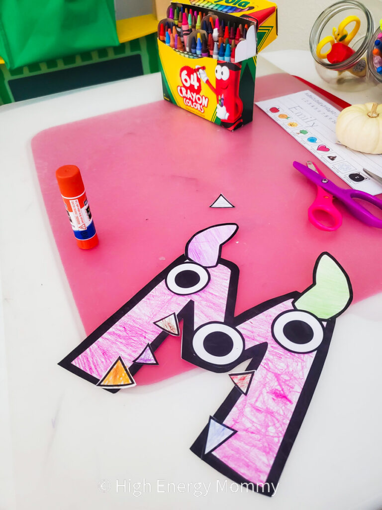 Craft with white paper cut into a letter M that was colored with pink and paper eyes, horns and teeth have been glued on to resemble a monster