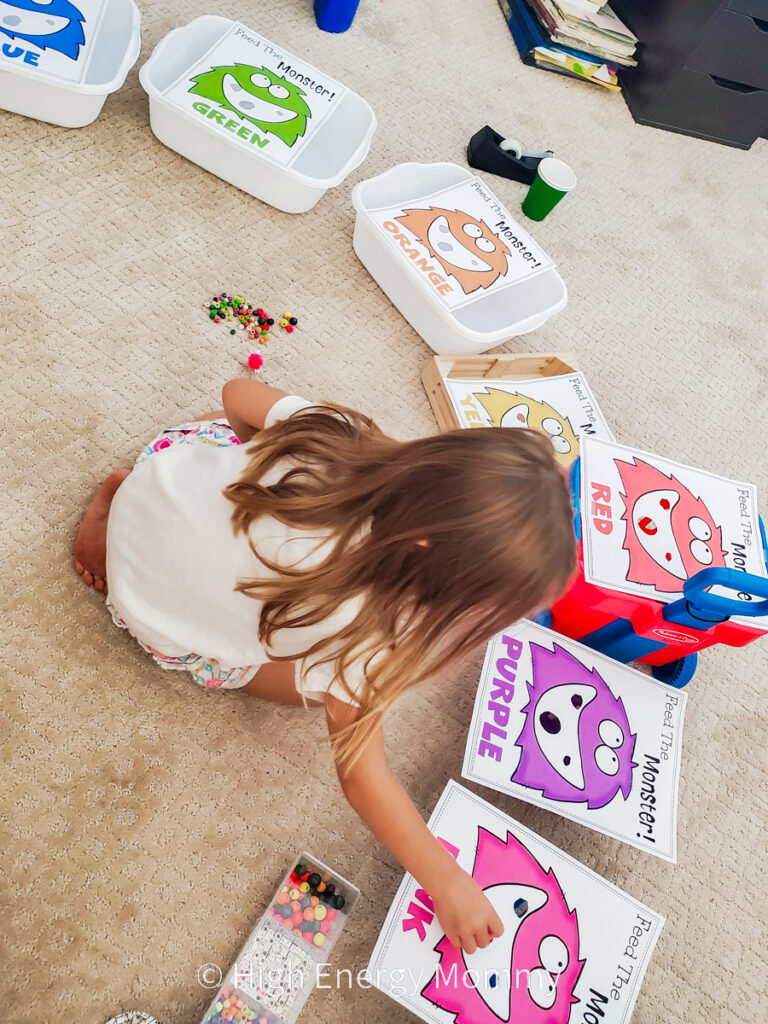 Side view of preschool age girl sitting on carpet with pages of different colored monster faces around her that are each taped to a small container. She is putting colored pom poms into the cut out monster mouths to color match