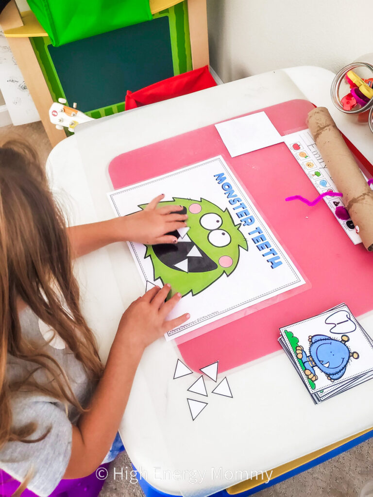 White piece of paper showing a cartoon monster face with large black mouth and toddler girl putting white triangles in the mouth like teeth