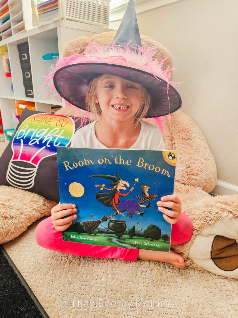 Little girl wearing a black witch hat with pink feather trim, holding room on the broom book 
