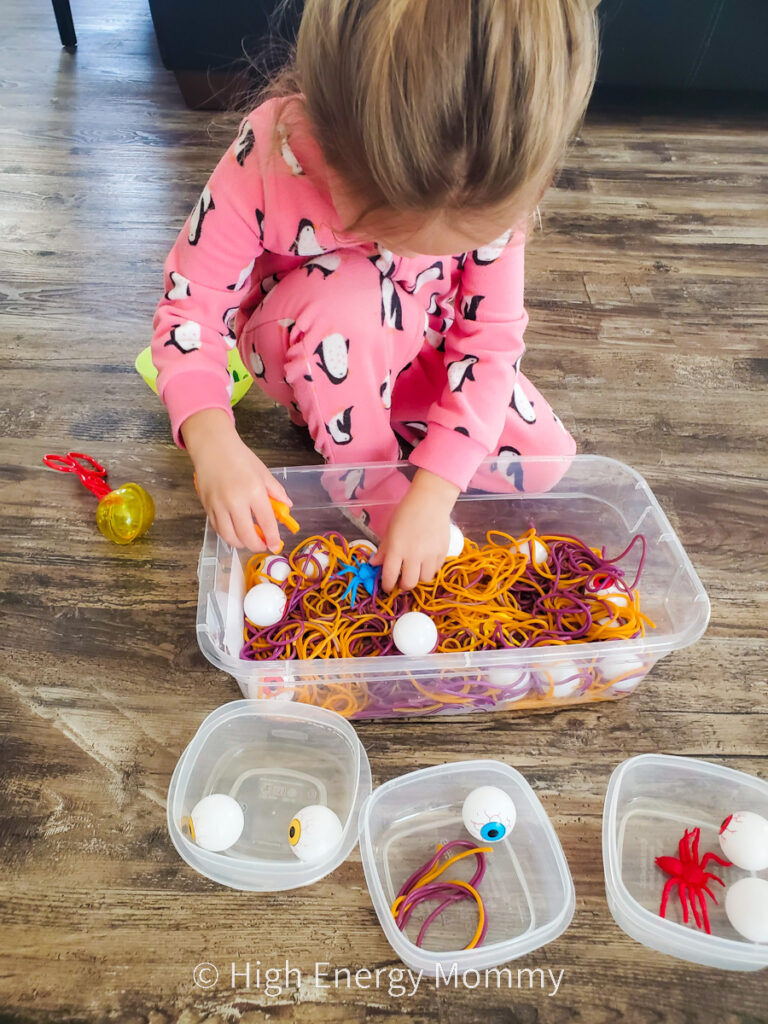 Toddler girl using hands in bin of cooked orange and purple colored noodles, plastic eyeballs and plastic spiders