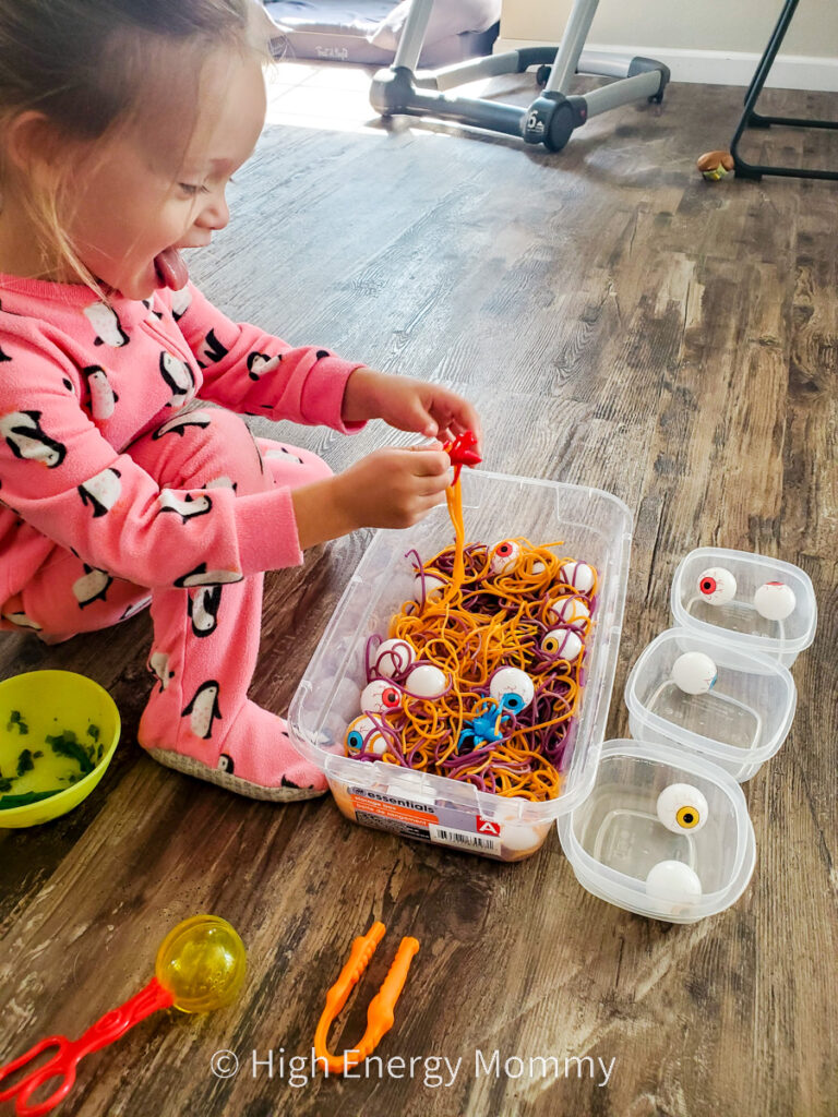 Toddler girl sticking tongue out, using hands in bin of orange and purple colored noodles, plastic eyeballs and plastic spiders