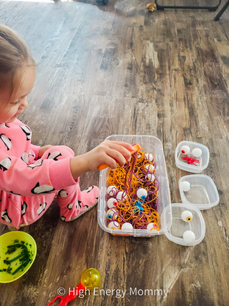 Toddler girl using plastic tweezers in bin of orange and purple colored noodles, plastic eyeballs and plastic spiders