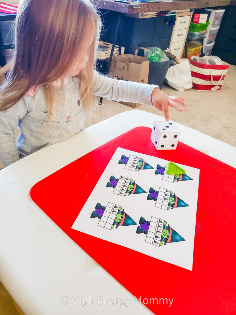 Preschool girl playing a dice game with a large die and math game mat of witch ten-frames from 1 to 6.