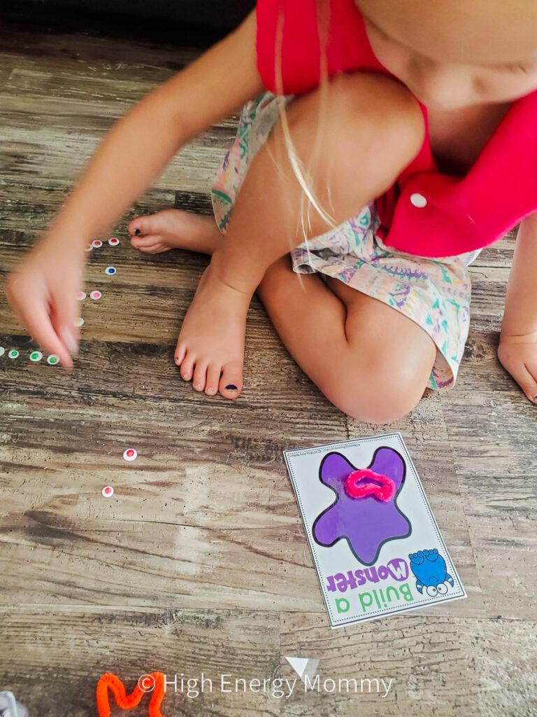 Toddler girl sitting on hardwood floor with a small piece of paper with a purple blob on it that she's putting googly eyes and pipe cleaners on to make a face