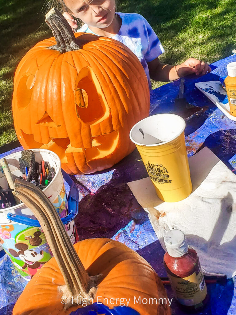 child painting the back side of a carved pumpkin on a table with paint and art supplies all around