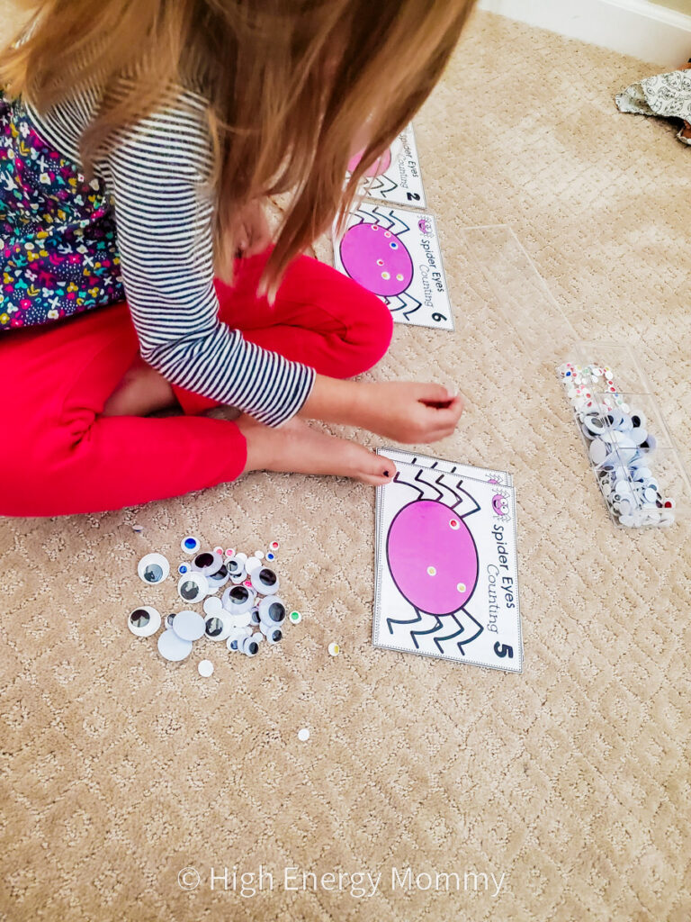 overhead view of toddler girl putting googly eyes onto a picture of a purple cartoon spider 