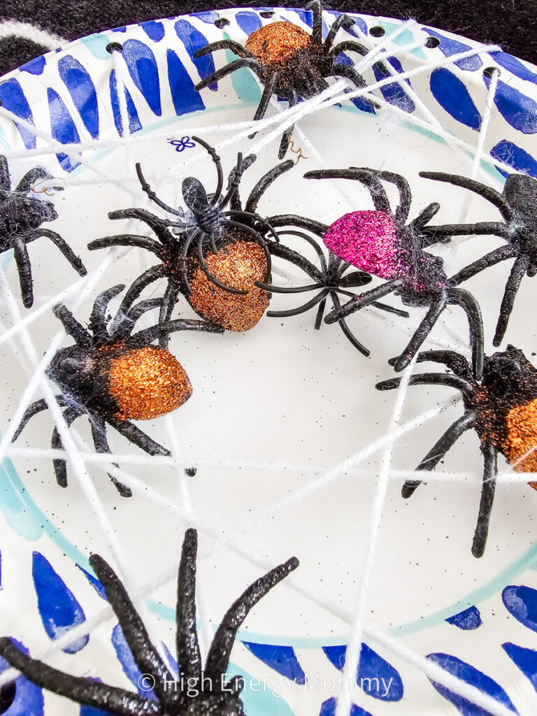 blue and white paper plate with white string attached in cross pattern like a spider web with purple, orange and black glitter plastic spiders