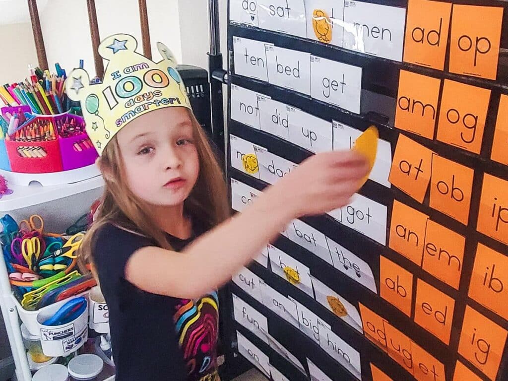 A child wearing a "100 days smarter" crown points at a word chart with various words and word endings, in a classroom setting with school supplies in the background.