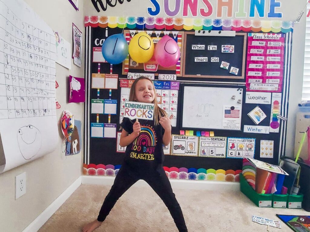 A child stands in a colorful classroom holding a sign that says "Kindergarten Rocks." The room is decorated with educational posters, balloons, and a calendar.