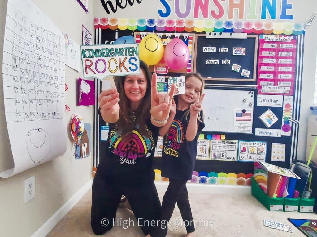 A child and woman pose in a colorful classroom holding a signs that say "Kindergarten Rocks" and "I am a smart cookie". The room is decorated with educational posters, balloons, and a calendar.