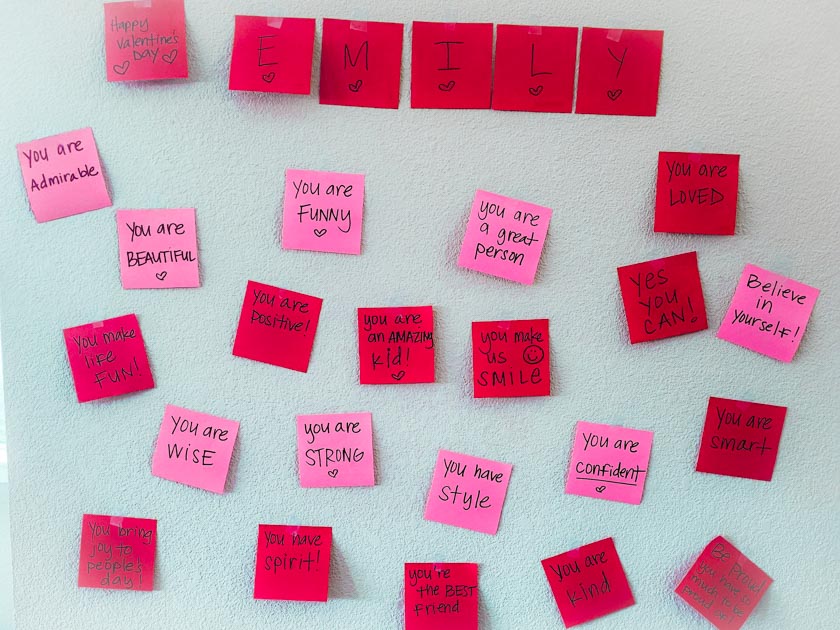 Pink and red Post It note papers stuck to a wall with a handwritten positive note or compliment on each paper.