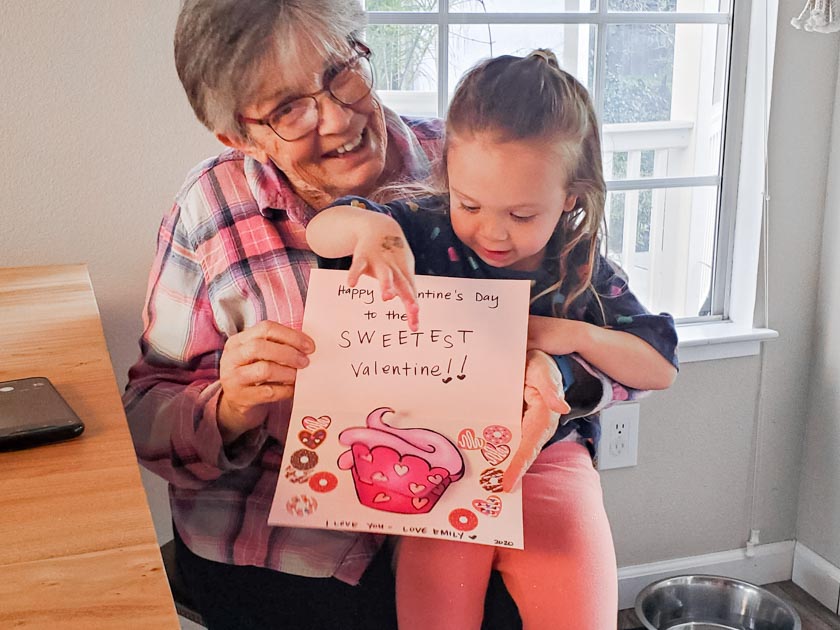 Grandmother sitting with toddler on her lap holding up a homemade valentine card with a pink paper cupcake popping out of the middle of the card. Card message says Happy Valentine's Day to the Sweetest Valentine.