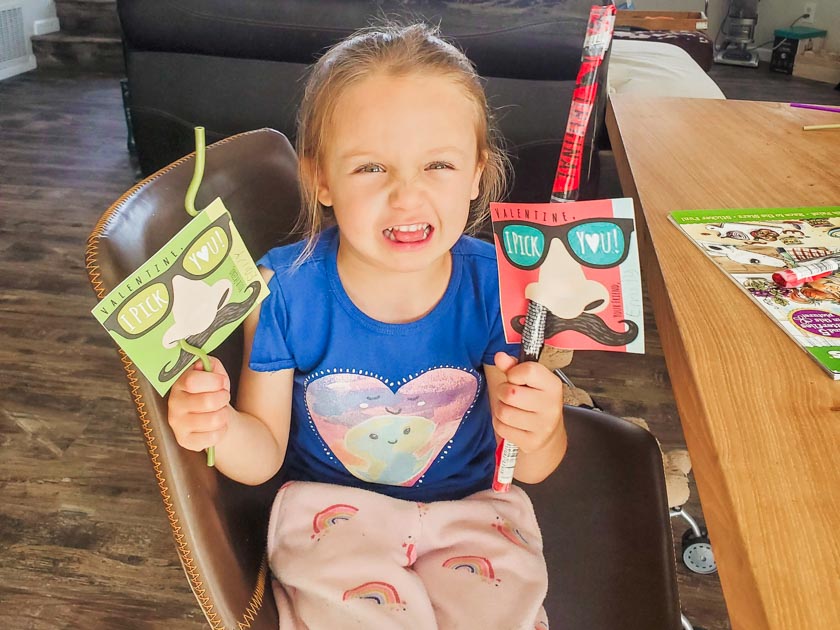 Little girl holding a plastic straw in each hand with valentine cards attached that have a large nose, mustache, and glasses and say Valentine, I pick you. The straw goes through the nostril of the card