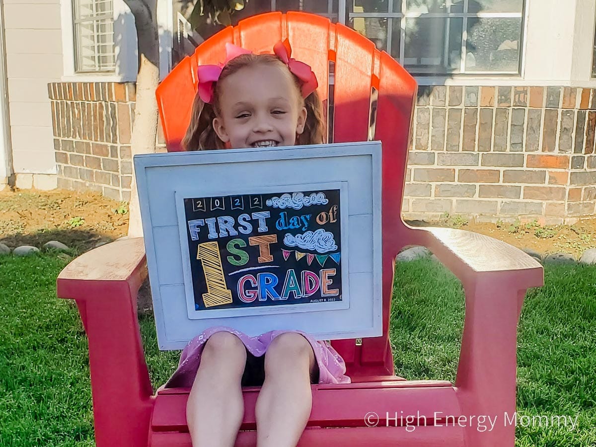 Young girl sitting outside on a red chair holding a sign that says First Day of 1st Grade