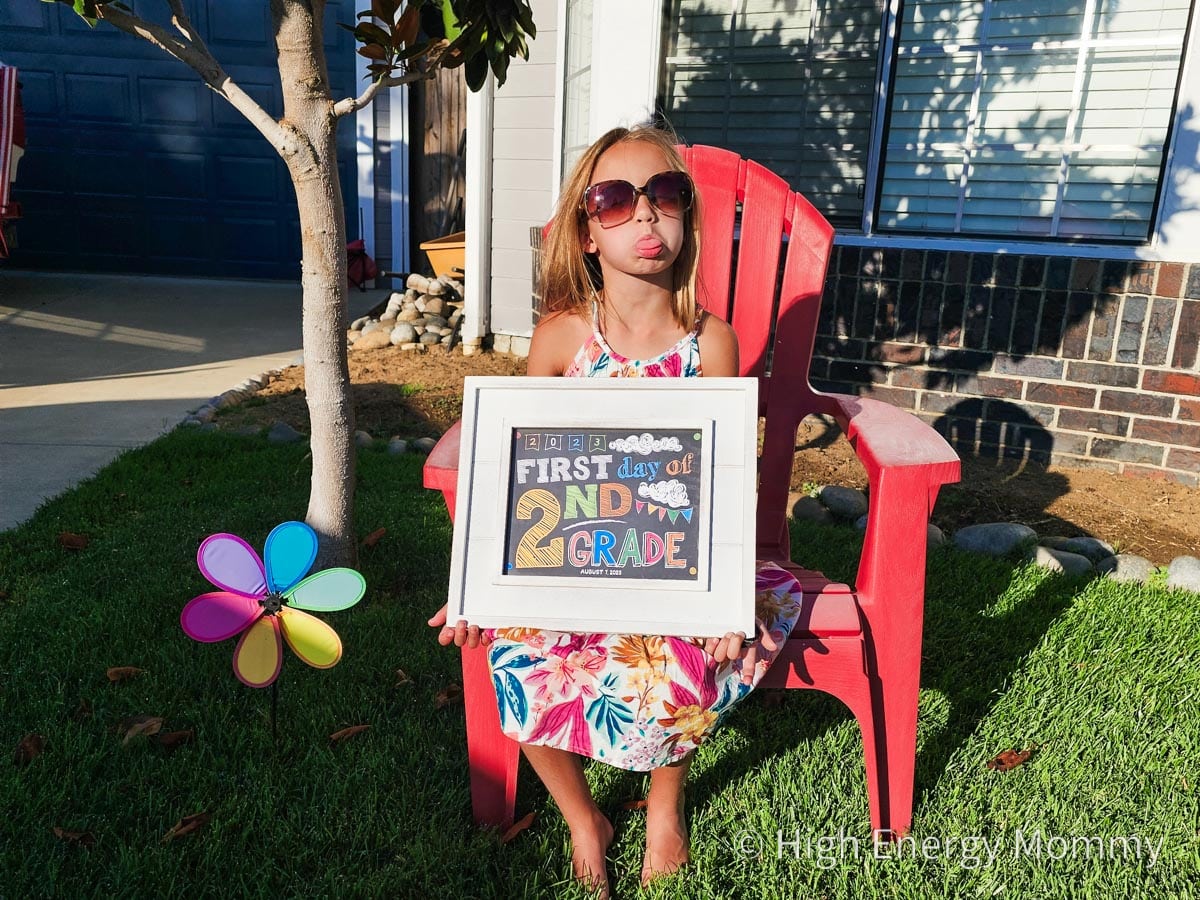 Young girl sitting outside on a red chair holding a first day of 2nd grade sign. The girl is wearing big sunglasses and sticking out her tongue.