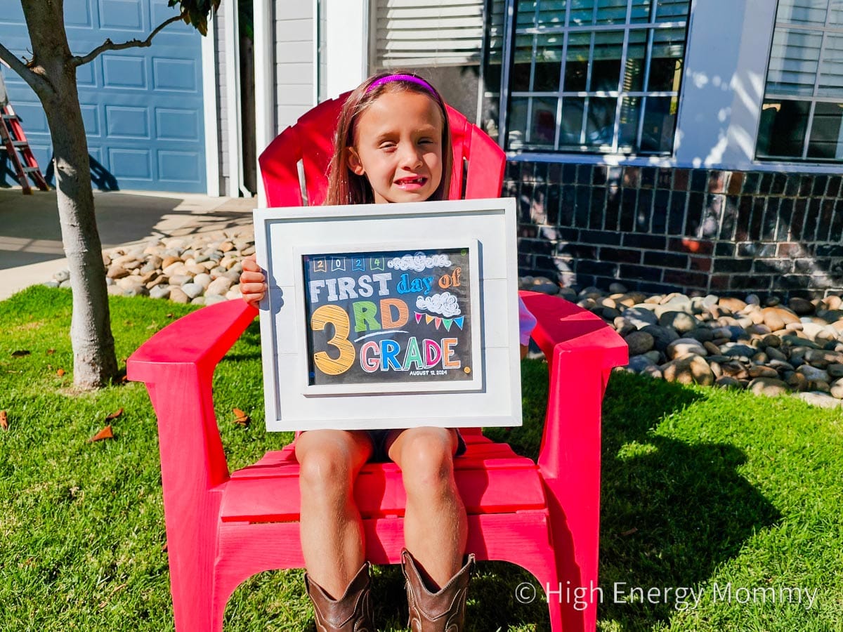 Young girl sitting outside on a red chair holding a first day of 3rd grade sign