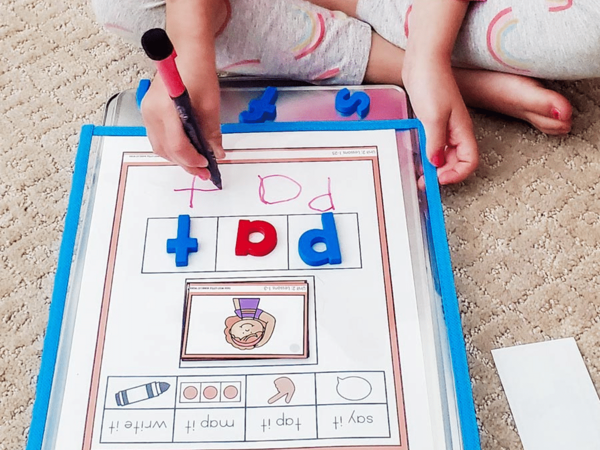 Child writing the word "Pat" on a dry erase mat with magnetic letters also spelling PAT
