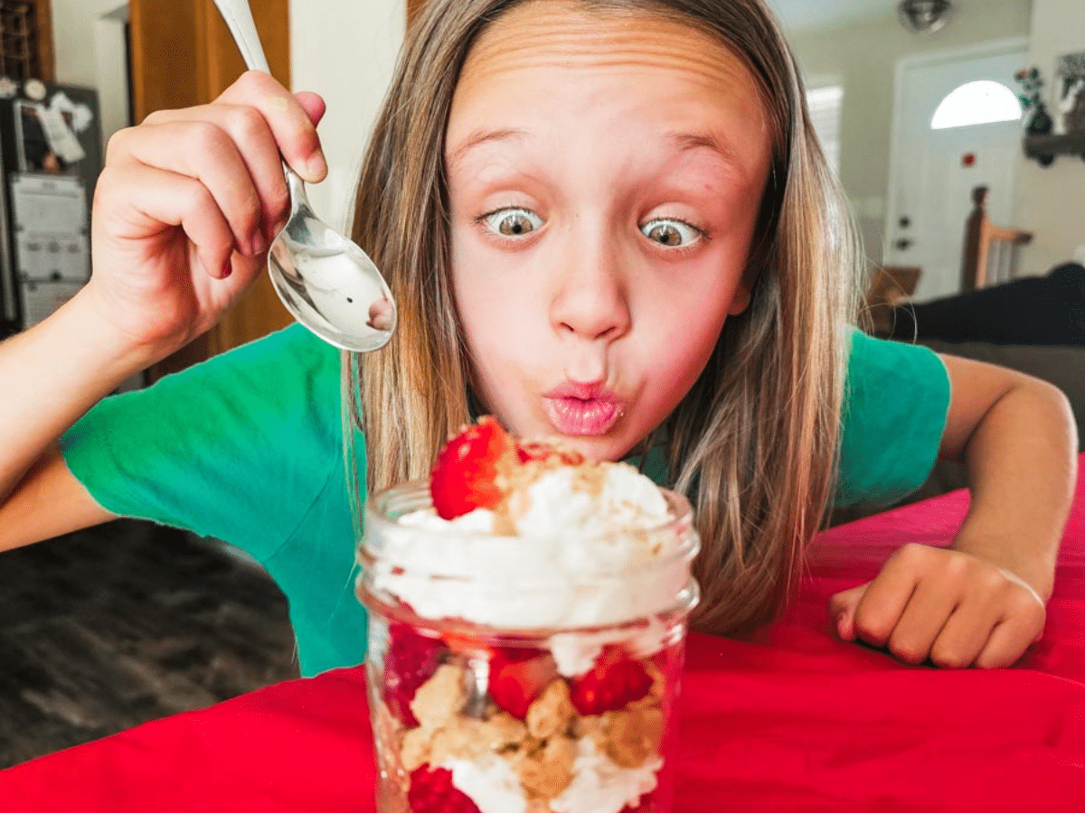 Young girl with an excited look on her face sitting. She is holding up a spoon behind a strawberry and whipped cream dessert