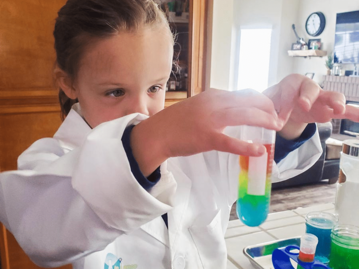 Little girl wearing a white lab coat holding up a beaker full of rainbow colored liquid