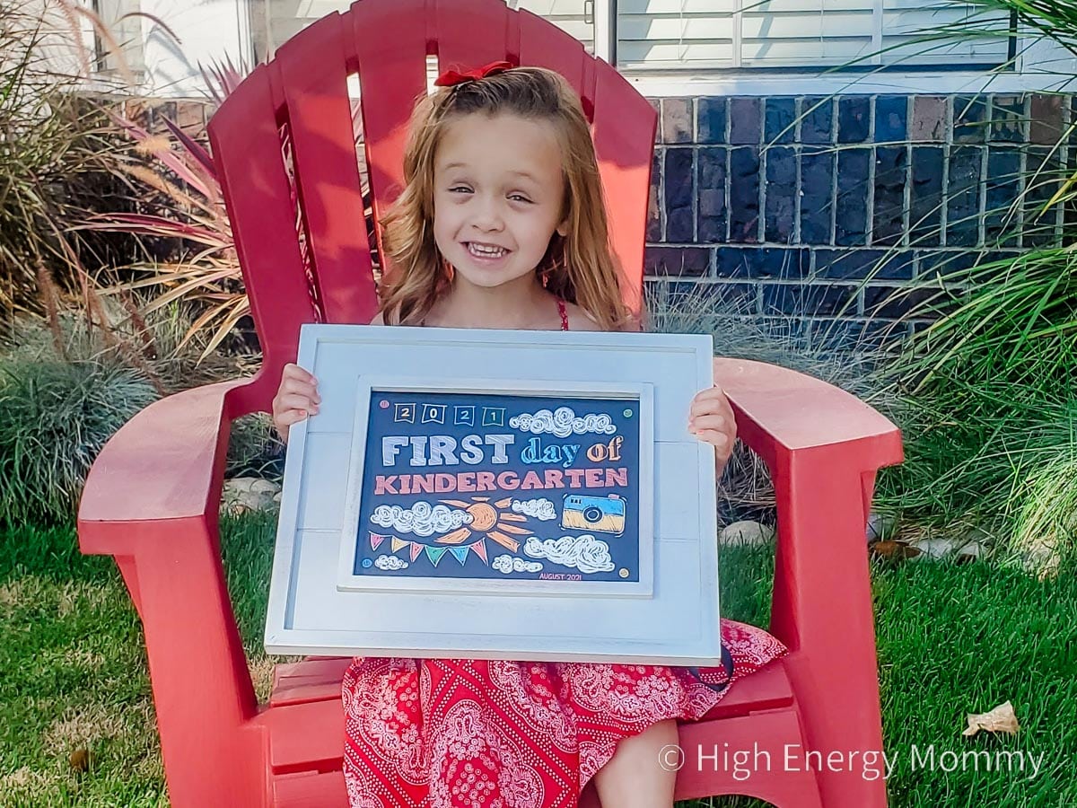 Young girl sitting outside on a red chair holding a first day of kindergarten sign