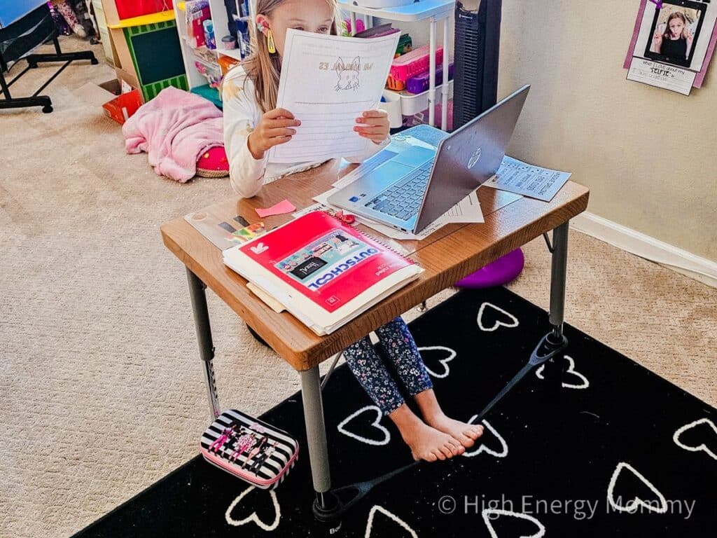 Child sitting at desk with laptop and reading a piece of paper with feet on desk band