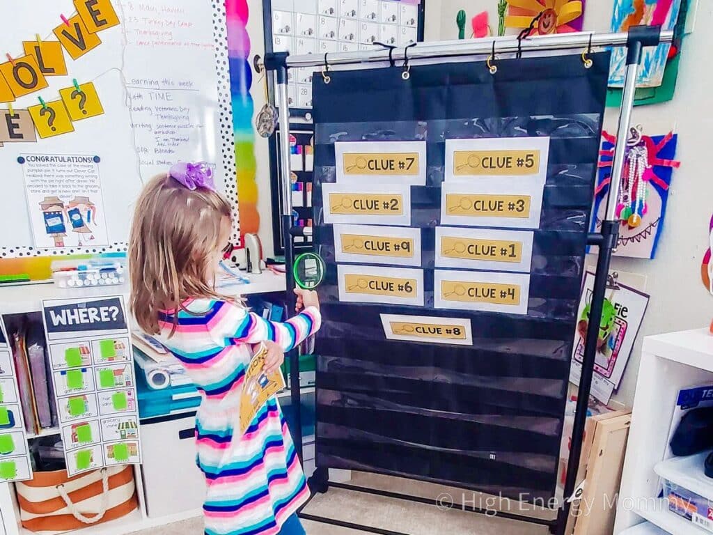 Child with a magnifying glass standing in front of a black pocket chart that has envelopes of clues in the pockets