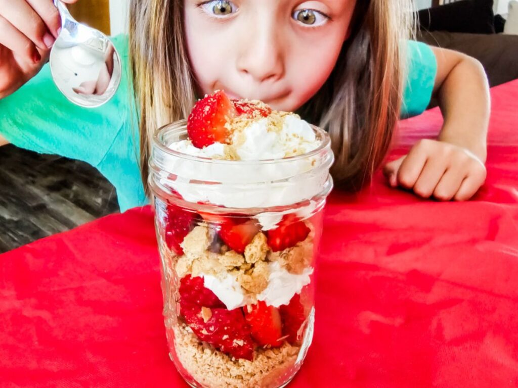 Child eagerly holding a spoon behind a strawberry layered mini mason jar dessert filled with fruit and whipped cream.